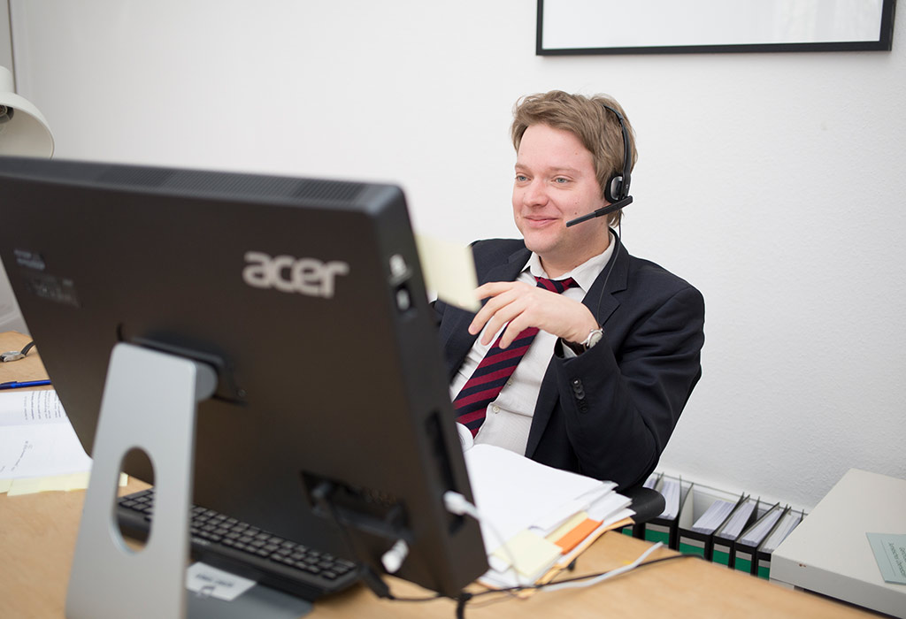 Ein Mann in Anzug lächelt am Schreibtisch, trägt ein Headset vor einem großen Computermonitor.